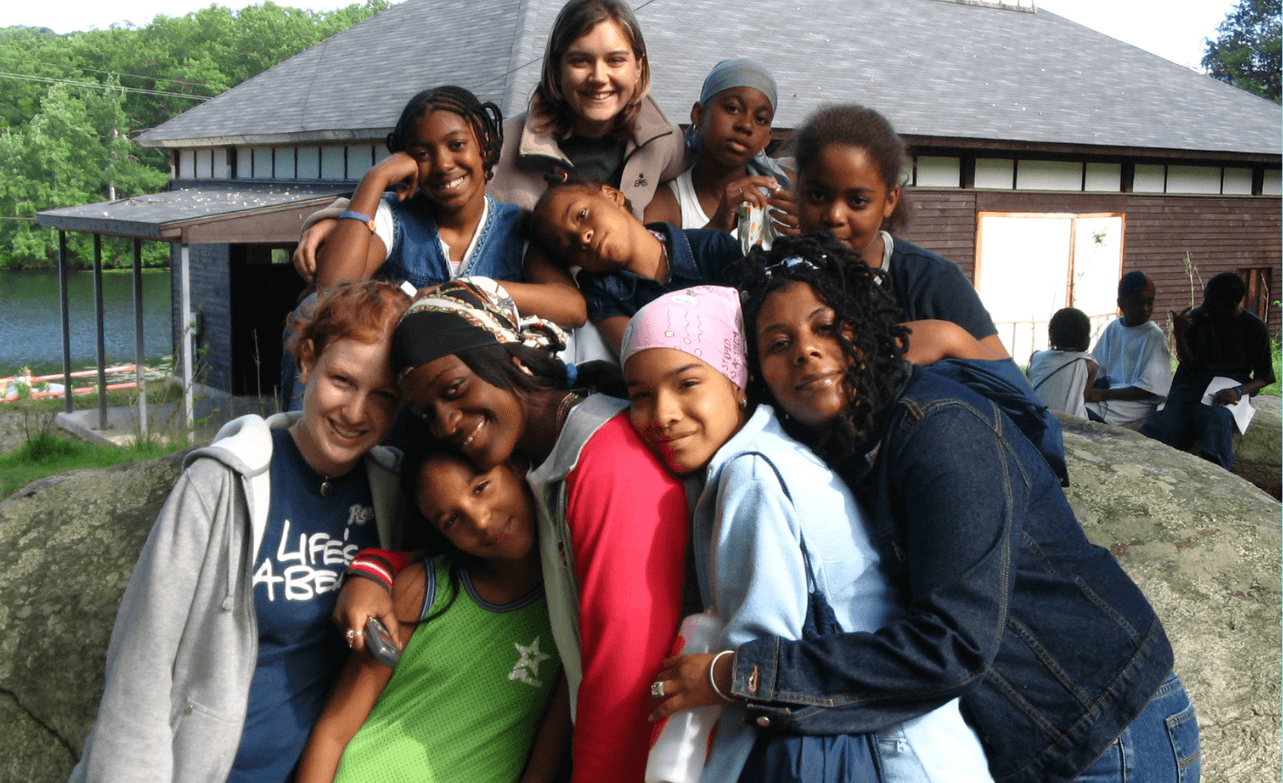 A photo from 2004 of 10 friends grouped together, hugging and smiling at camera. They are outside in a natural setting.